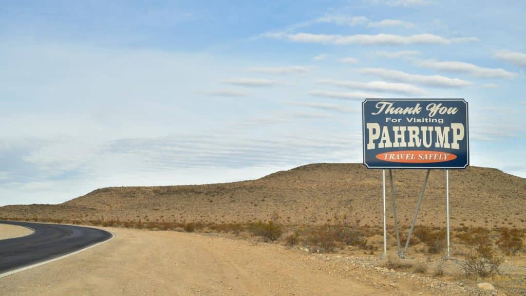 Roadside sign reading 'Thank You for Visiting Pahrump, Travel Safely' in a desert landscape with hills and cloudy sky.