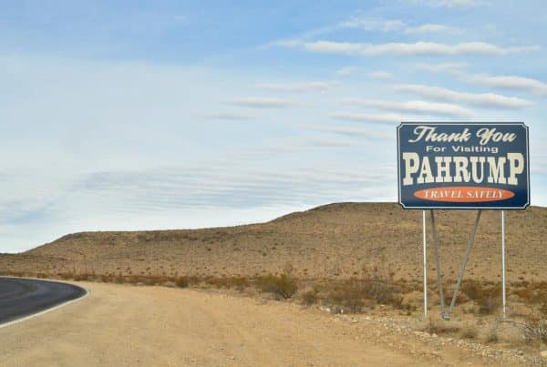 Roadside sign reading 'Thank You for Visiting Pahrump, Travel Safely' in a desert landscape with hills and cloudy sky.