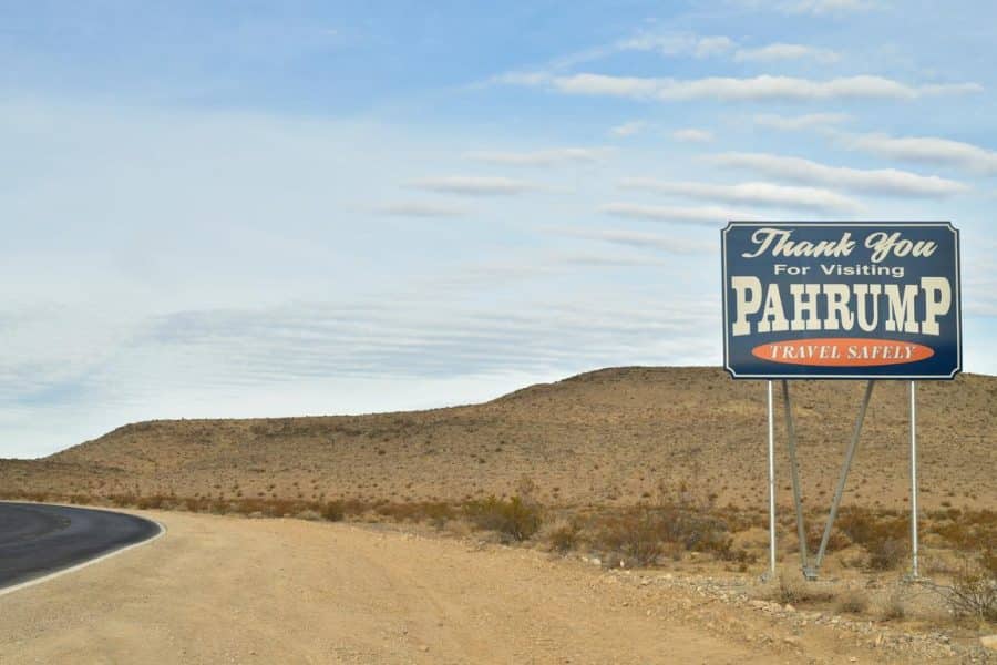 Roadside sign reading 'Thank You for Visiting Pahrump, Travel Safely' in a desert landscape with hills and cloudy sky.
