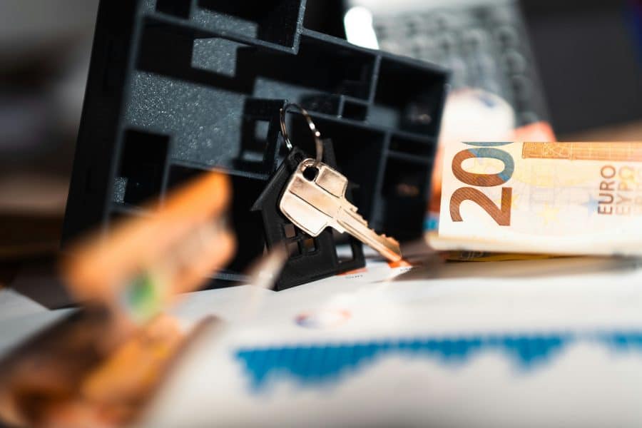 Close-up of house keys on a keychain with a house-shaped keyring, euro banknotes, and financial documents in the background.