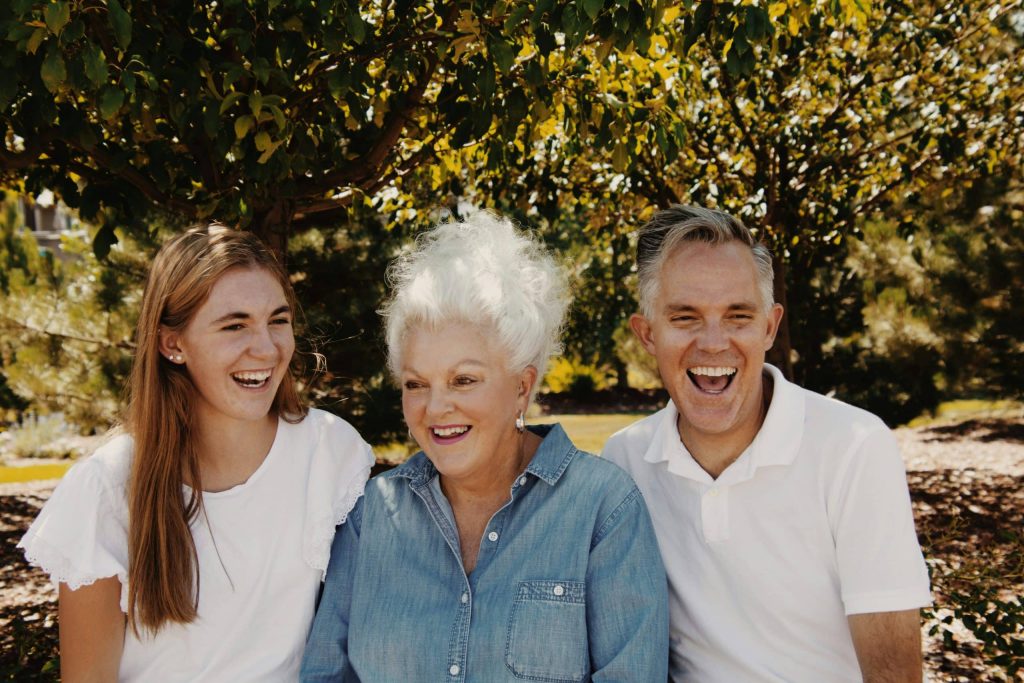 Three people outdoors sitting under trees, smiling and laughing together, including a young woman, an older woman with white hair, and a man.