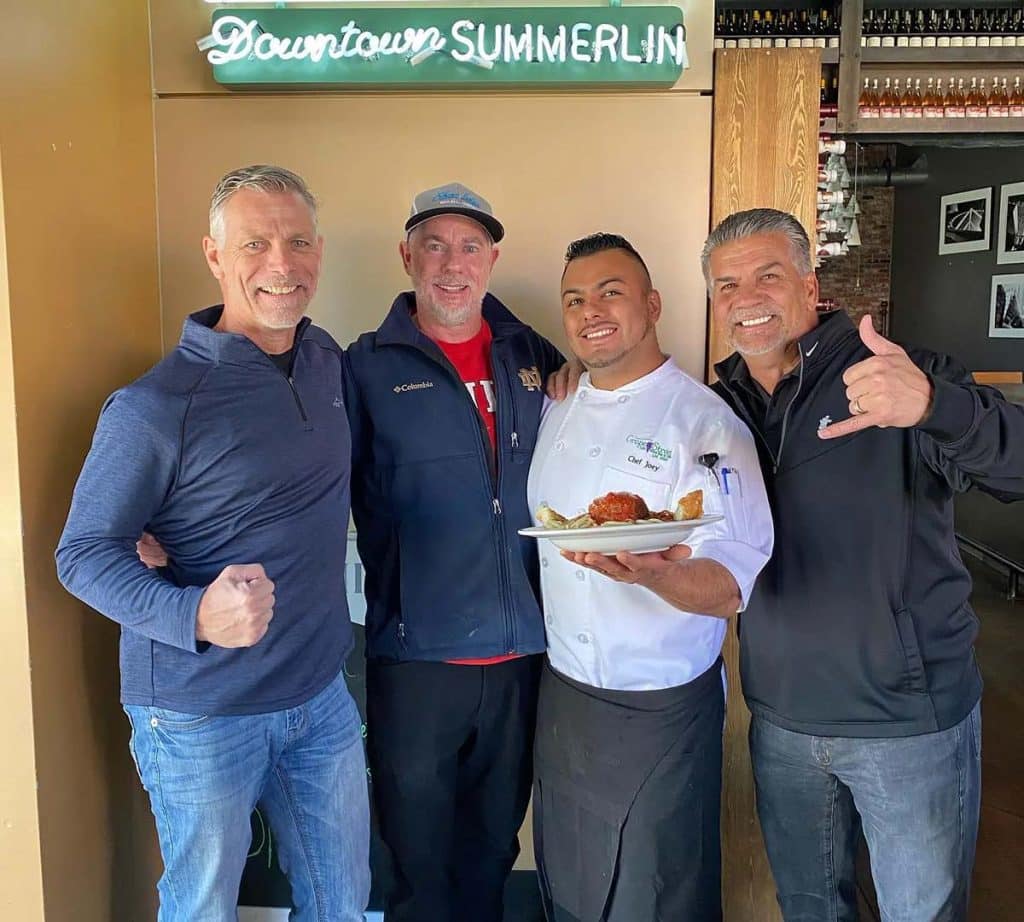 Four smiling men pose, one chef holding a plate of food, under a "Downtown SUMMERLIN" sign.
