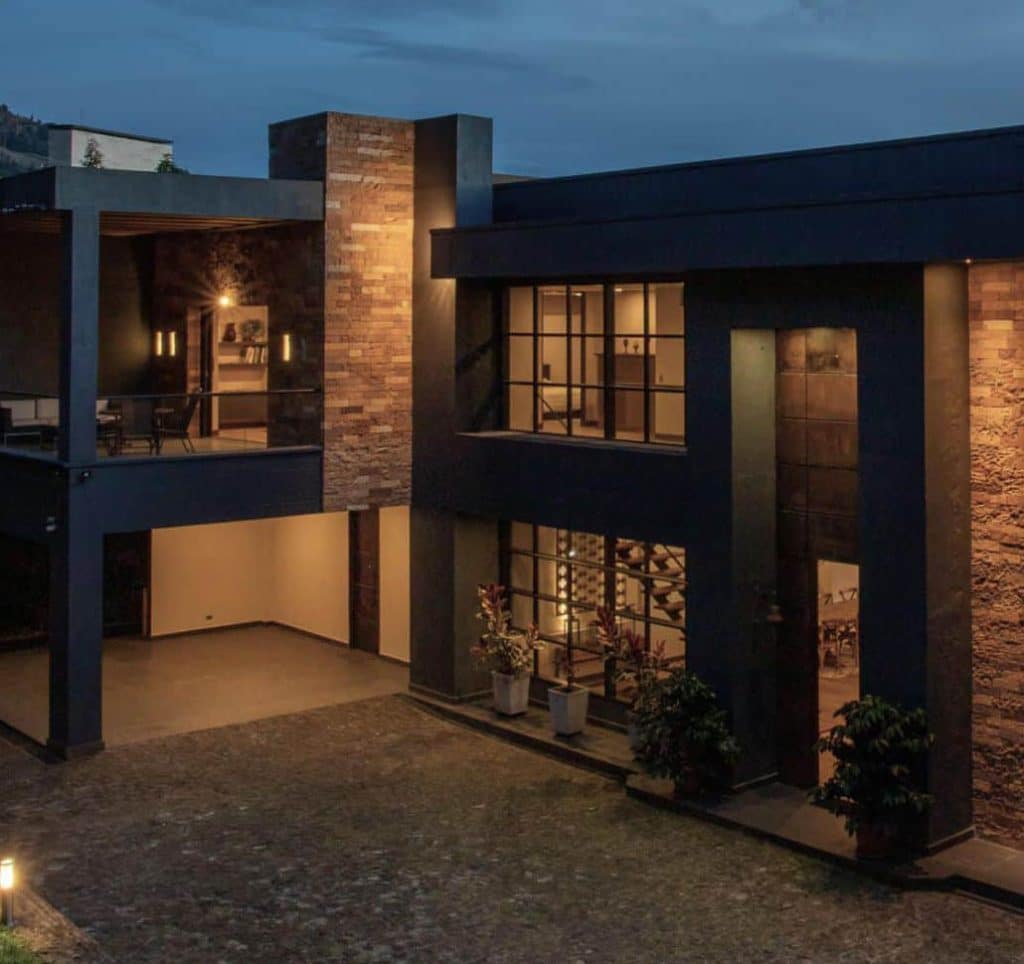 A modern, dark-colored building with brick accents and large windows, illuminated at dusk.