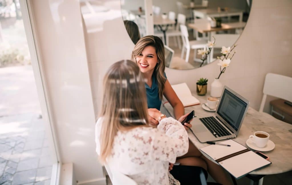 Two women sit at a cafe table with a laptop, coffee, and notebooks; one woman smiles at the other.