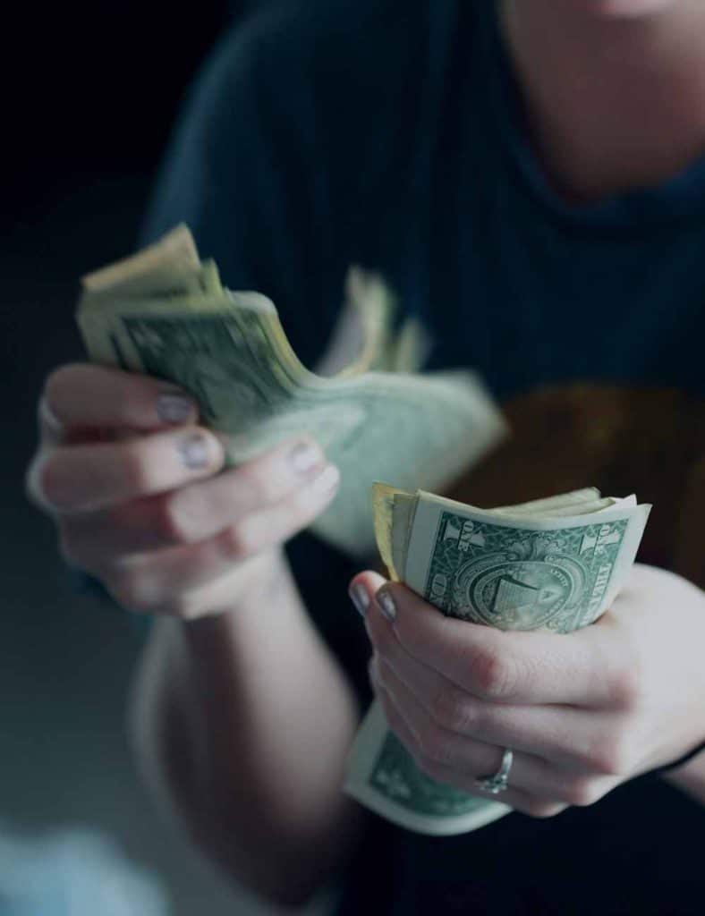A person with silver nail polish holds two wads of U.S. dollar bills, one with "ONE" visible.