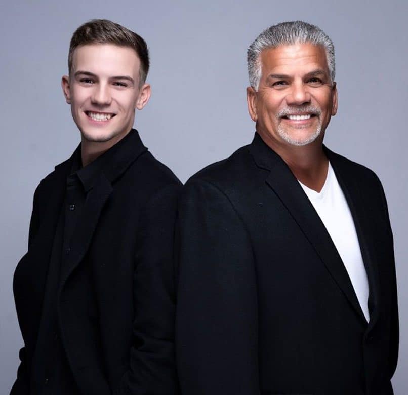 A young man and an older man with gray hair and a beard, both smiling and wearing black blazers.