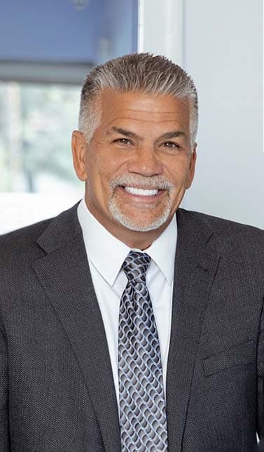 A smiling man with gray hair and a goatee wears a dark suit, white shirt, and patterned tie.