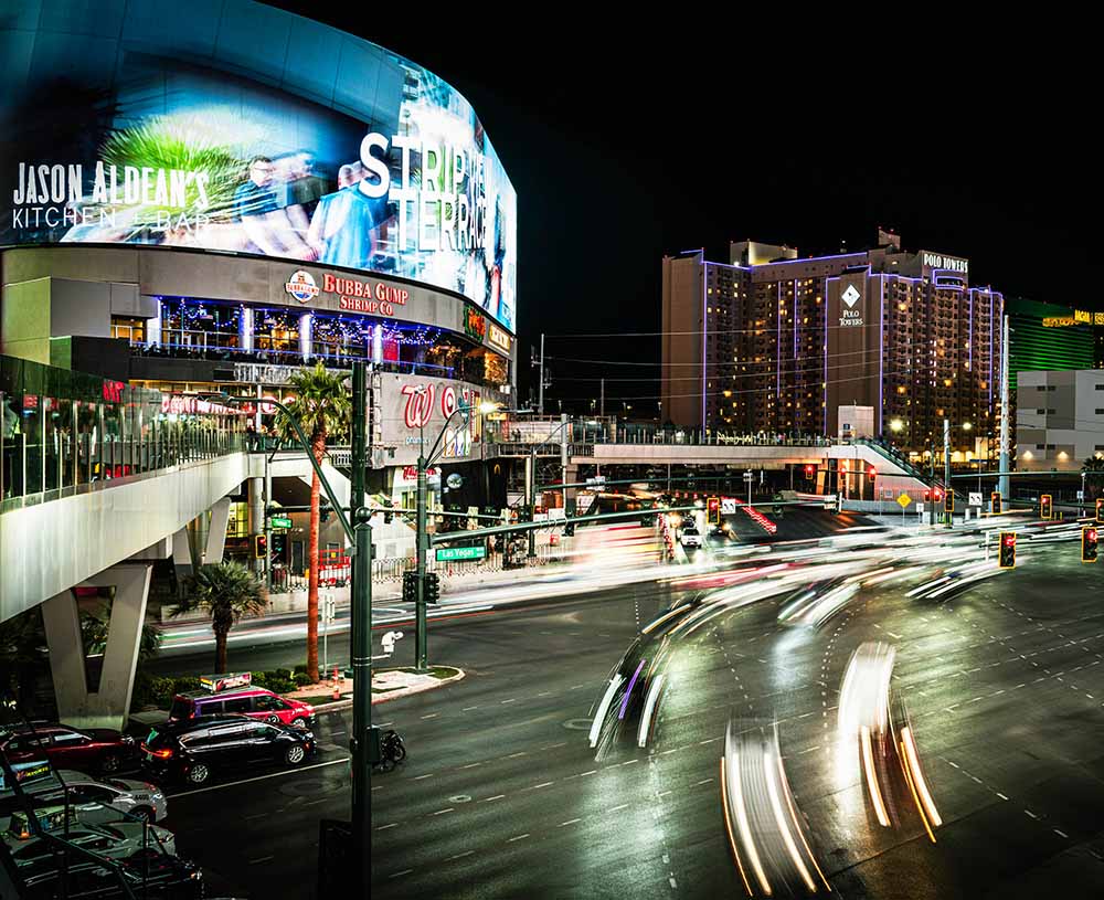 A vibrant night scene shows a curved building with "JASON ALDEAN'S KITCHEN + BAR" and "STRIP TERRACE" signs, a hotel, and blurred car lights on a street.