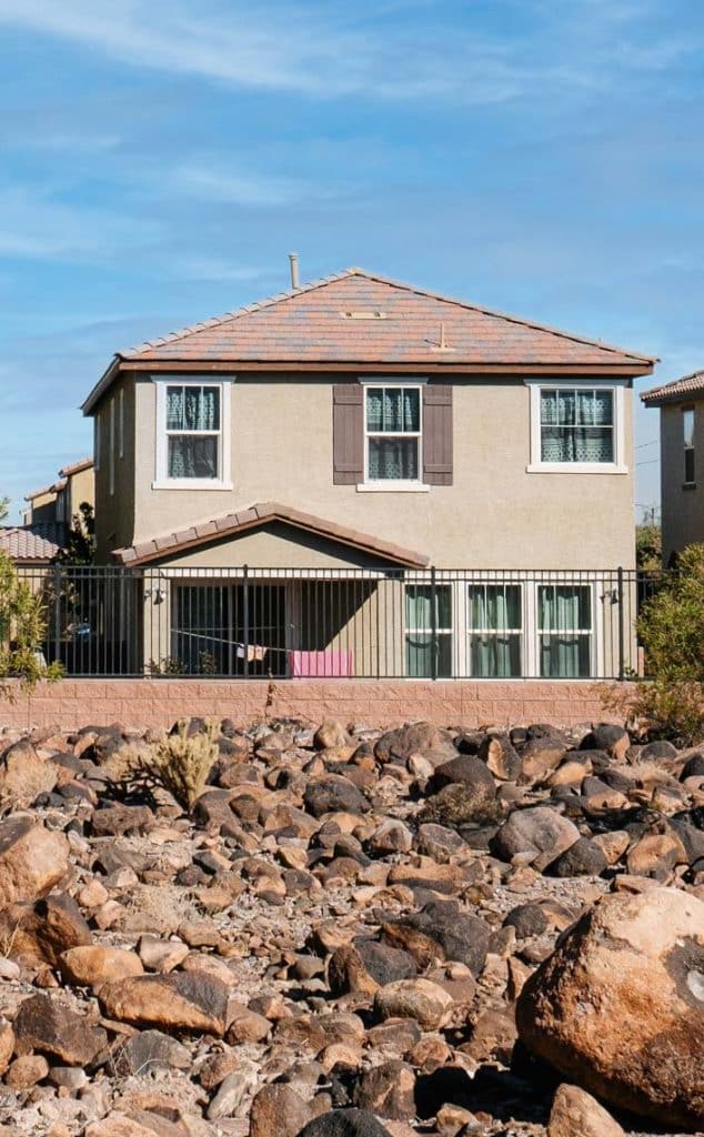A two-story stucco house with a tiled roof sits behind a black fence and a field of rocks.