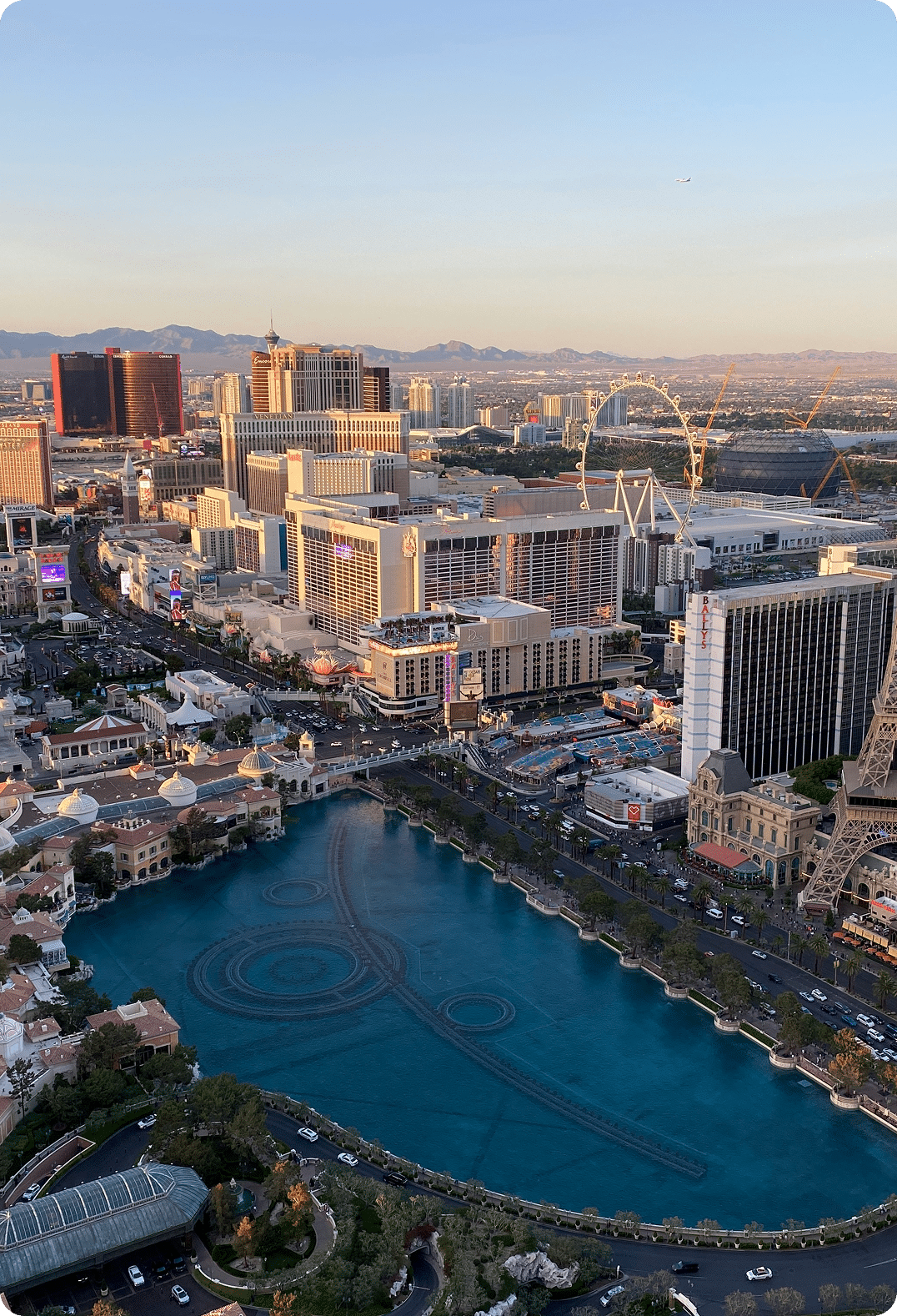 An aerial view of the Las Vegas Strip, with hotels, a large lake, and mountains in the distance.