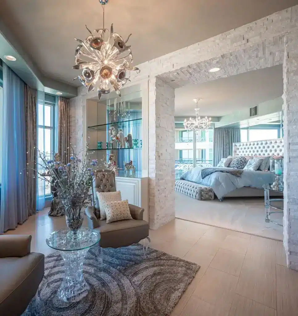 A luxurious living area with a unique chandelier, leading to a bedroom with a tufted headboard and blue bedding.