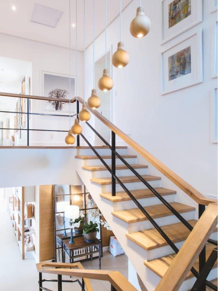 A modern staircase with light wood treads, black railings, and hanging globe lights descends into a bright room.